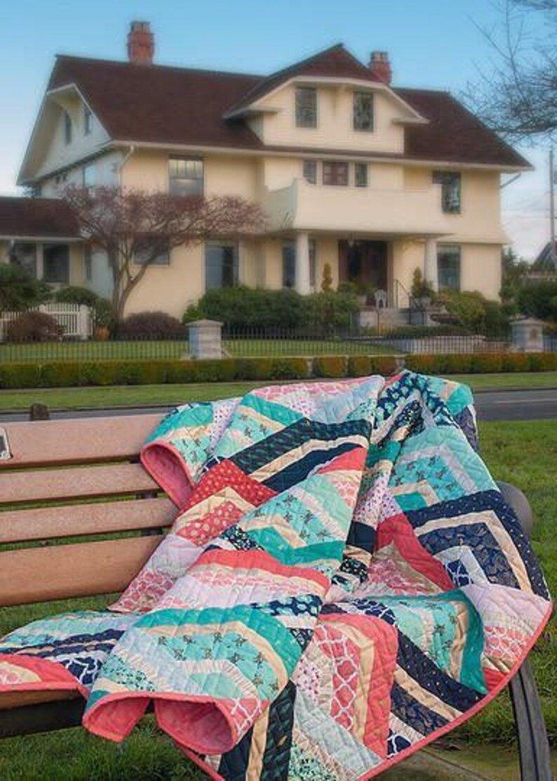 K&ouml;nnte beinhalten: Eine bunte Patchwork-Decke mit einem Chevron-Muster in Rosa-, Blau-, Gr&uuml;n- und Wei&szlig;t&ouml;nen liegt &uuml;ber einer Holzbank. Die Decke ist gefaltet und hat eine weiche, strukturierte Oberfl&auml;che.