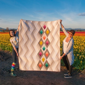 Puede incluir: Dos personas sostienen una gran colcha con un patr&oacute;n geom&eacute;trico de diamantes coloridos sobre un fondo blanco. La colcha est&aacute; frente a un campo de tulipes amarillas.
