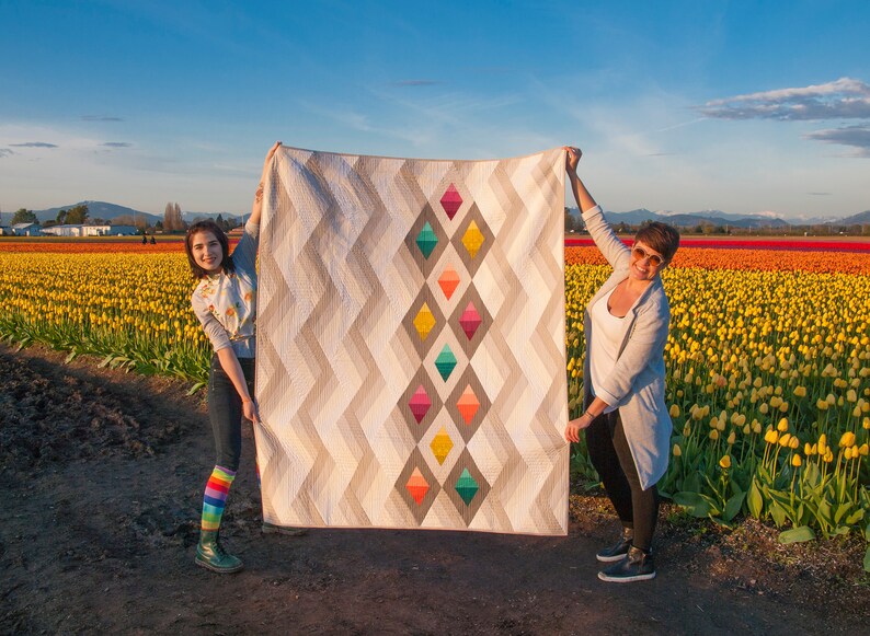 Puede incluir: Dos personas sostienen una gran colcha con un patr&oacute;n geom&eacute;trico de diamantes coloridos sobre un fondo blanco. La colcha est&aacute; frente a un campo de tulipes amarillas.