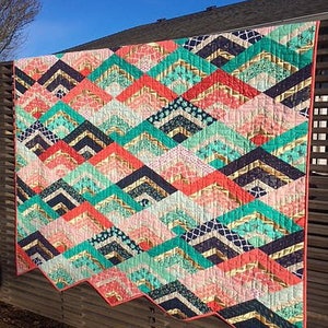 K&ouml;nnte beinhalten: Ein farbenfroher Quilt mit einem Chevron-Muster in den Farben Rosa, T&uuml;rkis, Gold und Schwarz. Der Quilt h&auml;ngt an einem Holzzaun.