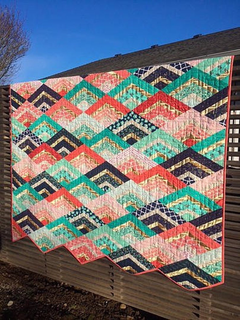 K&ouml;nnte beinhalten: Ein farbenfroher Quilt mit einem Chevron-Muster in den Farben Rosa, T&uuml;rkis, Gold und Schwarz. Der Quilt h&auml;ngt an einem Holzzaun.