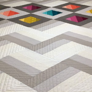 Puede incluir: Una colcha blanca y gris con un patr&oacute;n de chevron. La colcha tiene un patr&oacute;n de diamante en el centro con cuadrados coloridos en cada diamante.