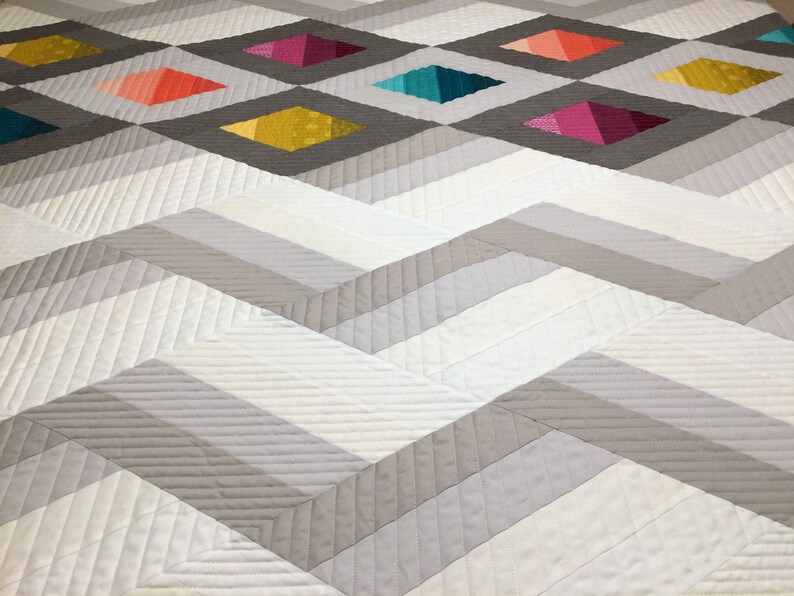 Puede incluir: Una colcha blanca y gris con un patr&oacute;n de chevron. La colcha tiene un patr&oacute;n de diamante en el centro con cuadrados coloridos en cada diamante.