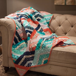 K&ouml;nnte beinhalten: Eine bunte Patchwork-Decke mit einem Chevron-Muster in den Farben T&uuml;rkis, Koralle, Wei&szlig; und Marineblau. Die Decke liegt &uuml;ber einem hellbeigen Sofa.