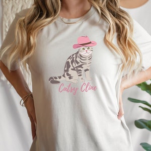 May include: A white t-shirt with a graphic of a cat wearing a pink cowboy hat. The text "Cattsy Cline" is printed below the cat.