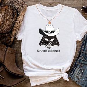 Peut inclure: T-shirt blanc avec un motif noir et blanc de Dark Vador portant un chapeau de cowboy. Le texte "Darth Brooks" est imprimé en dessous. Le t-shirt est présenté avec un collier, des lunettes de soleil et un short en jean.