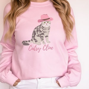 May include: Pink sweatshirt with a graphic of a cat wearing a pink cowboy hat. The text "Catsy Cline" is printed below the cat.