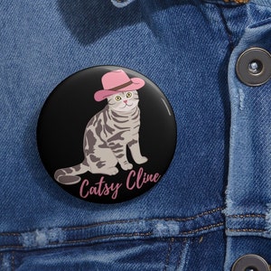 May include: A round black button featuring a cartoon cat wearing a pink cowboy hat. The cat has gray and white stripes and is sitting. The text "Catsy Cline" is written in pink cursive below the cat.