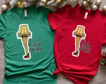 May include: Two t-shirts with a Christmas-themed design. The design features a leg wearing a fishnet stocking and a high heel, with a lampshade on top. The text on the shirts reads "It's a Major Award". One shirt is green and the other is red.