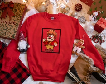 May include: Red Christmas sweatshirt with a graphic of a cartoon character with red hair and a yellow shirt. The character is in a black frame and has a grumpy expression. The sweatshirt is on a white surface with Christmas decorations.