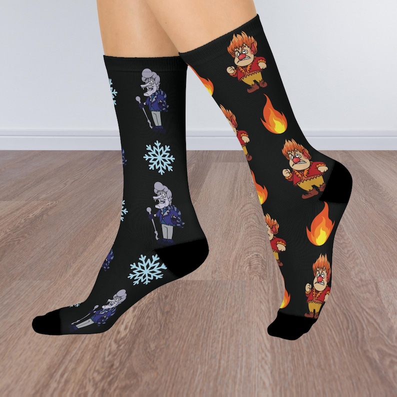 May include: A pair of black crew socks featuring mismatched designs. One sock displays a repeating pattern of a grey-haired character with a cane and light blue snowflakes. The other sock features a character with flaming hair and fire icons.