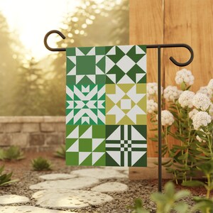 May include: A green and white geometric pattern garden flag. The flag features a patchwork design with various shapes and shades of green. The flag is displayed on a black metal stand in a garden setting.