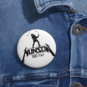 May include: A white button with a black silhouette of a person playing a guitar, the text "MUNSON 1986 Tour" and a few bats flying above the person.