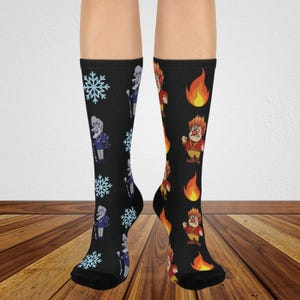 May include: A pair of black crew socks featuring a mismatched design. The left sock displays light blue snowflakes and a cartoon character with white hair. The right sock features orange and yellow flames and a cartoon character with red hair.