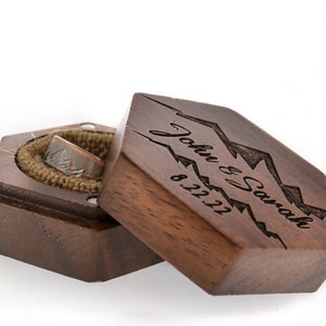 May include: A wooden ring box with a hexagonal shape. The lid is open and shows a ring inside. The lid is engraved with the text "John & Sarah 8.12.21" and a mountain range design.