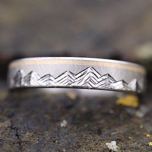 May include: A silver wedding band with a gold inlay and a mountain range design.