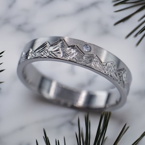 May include: A silver band with a mountain range design and a single diamond set in the center.
