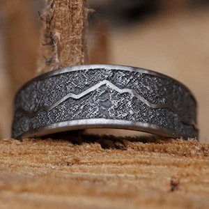 May include: A silver ring with a textured surface and a mountain range design.
