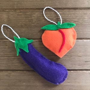 May include: Two felt ornaments: a purple eggplant and an orange peach. The eggplant has a green stem and a white string for hanging. The peach has a green stem and a red line down the side, also with a white string for hanging. The ornaments are on a wooden surface.
