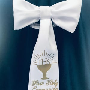 May include: White satin bow with a long, flowing tail. The tail features a gold chalice design with the text "First Holy Communion". The bow is attached to a dark blue surface.