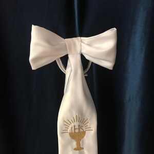 May include: White satin bow and stole with gold embroidery of a chalice and the letters IHS. The stole is attached to the bow with a thin white cord. The background is a dark blue fabric.