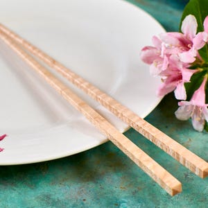 May include: A pair of wooden chopsticks on a white plate with pink flowers. The chopsticks are made of light wood with a natural grain pattern.