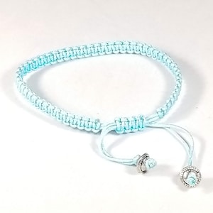 May include: A light blue braided bracelet with silver metal accents. The bracelet has a sliding knot for adjusting the fit.