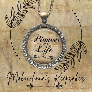 May include: A round pendant necklace with the words "Pioneer Life" in elegant script. The pendant is surrounded by a ring of clear crystals and hangs from a silver chain. The background is a textured, sepia-toned surface with decorative leaf illustrations.