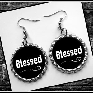 May include: A pair of black and silver earrings. Each earring features a black circular design with the word "Blessed" in white script. The earrings are set against a white background and a dark wood grain surface.