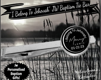May include: A silver tie bar with a black circular charm that reads "I Belong To Jehovah! Manny LEWIS 01-21-23". The tie bar is photographed against a black and white background of tall grass and a lake.