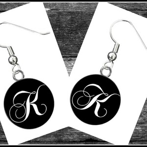 May include: A pair of silver dangle earrings with black round charms featuring a white cursive letter K.