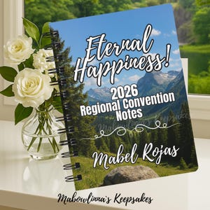 May include: A notebook with a mountain landscape cover, spiral-bound. The cover features the text "Eternal Happiness! 2026 Regional Convention Notes", the name "Mabel Rojas", and "Mabowlivina's Keepsakes". A vase of white roses is visible to the left.