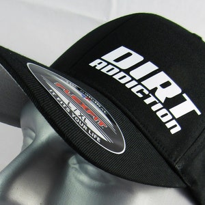 May include: A black baseball cap with a white logo that says "Dirt Addiction". The cap has a grey underbill and a Flexfit label.