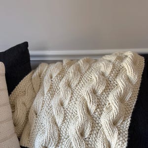 Puede incluir: Una manta de punto de color crema con un patrón de punto trenzado. La manta está colocada sobre un sofá negro.