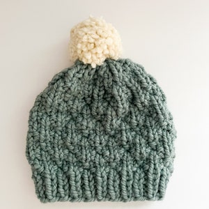 May include: A sage green knitted beanie with a white pom pom on top.