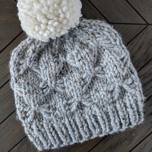 May include: A gray knitted beanie with a white pom pom on top. The beanie has a cable knit pattern.