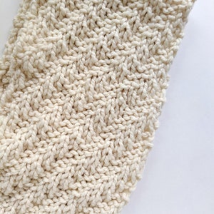 May include: A close-up of a cream-colored knitted fabric with a textured pattern. The fabric is soft and has a subtle sheen.