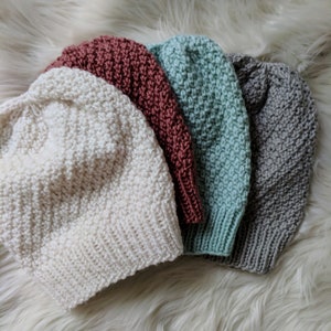 May include: Four knitted hats in different colors: white, light brown, light green, and rust. The hats are stacked on top of each other on a white fluffy surface.
