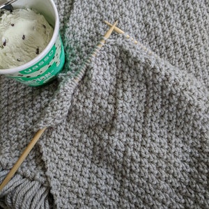K&ouml;nnte beinhalten: Eine graue Strickdecke mit Fransen an den R&auml;ndern. Die Decke wird mit Holzstricknadeln gestrickt.