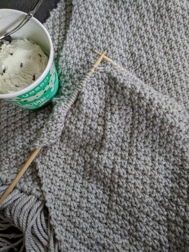 K&ouml;nnte beinhalten: Eine graue Strickdecke mit Fransen an den R&auml;ndern. Die Decke wird mit Holzstricknadeln gestrickt.