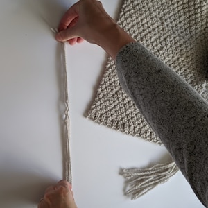 K&ouml;nnte beinhalten: Nahaufnahme einer Hand, die einen Knoten in eine lange, cremefarbene Schnur bindet. Die Schnur h&auml;ngt an einer beigefarbenen geh&auml;kelten Wandbeh&auml;nge mit Fransen.