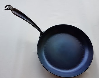 May include: A black cast iron skillet with a long handle. The handle has a unique loop design at the end. The skillet has a blue-grey finish.