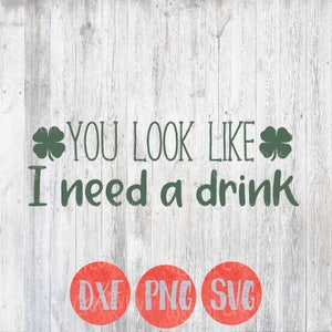 Puede incluir: Diseños de trébol verde sobre un fondo de madera blanca con el texto "YOU LOOK LIKE I need a drink". El texto está en verde. Debajo del texto hay tres círculos rojos con el texto "DXF", "PNG" y "SVG" en blanco.