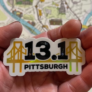 May include: A rectangular sticker with the text "13.1 PITTSBURGH" in black and gold. The design includes gold bridge illustrations. The sticker is held in a hand, with a map visible in the background.