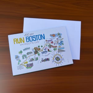 May include: A white card with a colorful map of Boston, Massachusetts, featuring the text "RUN BOSTON PATRIOT'S DAY" in blue and yellow. The card is accompanied by a white envelope, set against a wooden surface.