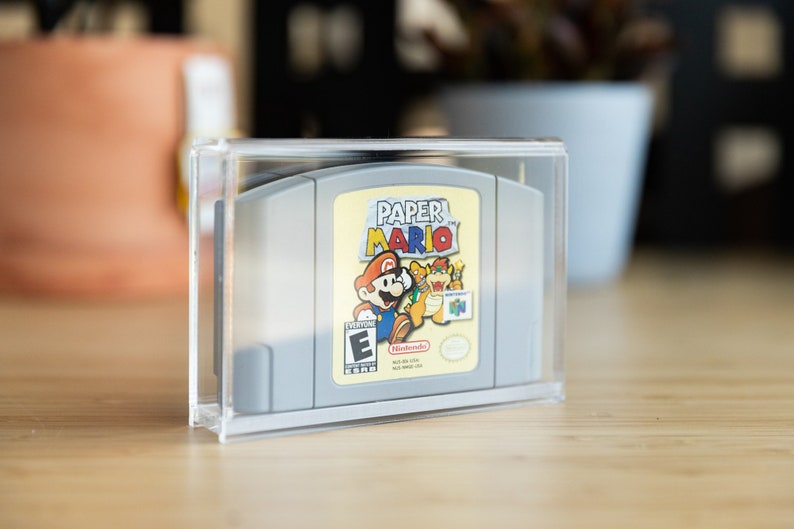 May include: A gray Nintendo 64 game cartridge for "Paper Mario" in a clear acrylic case. The game label features Mario, a yellow dragon, and the game title. The case is on a wooden surface, with blurred background elements.