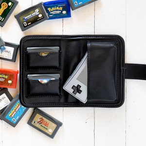 May include: Black leather Game Boy Advance cartridge case with a Game Boy Advance and several game cartridges. The case has pockets for cartridges and a snap closure. Game cartridges are scattered around the case.