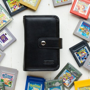May include: A black leather Game Boy Advance game cartridge case with a snap closure. The case is surrounded by a variety of Game Boy Advance game cartridges, including Pokemon, Zelda, and Mario games.