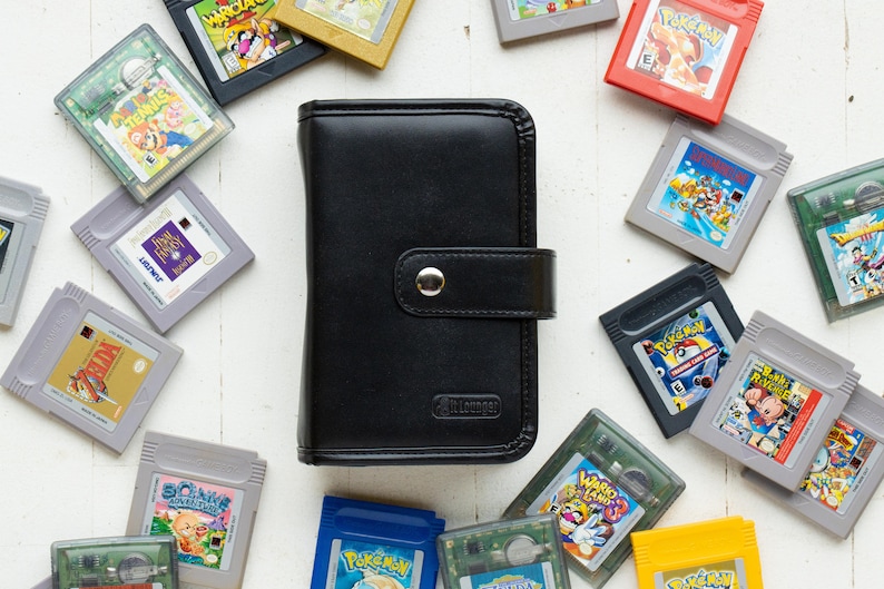 May include: A black leather Game Boy Advance game cartridge case with a snap closure. The case is surrounded by a variety of Game Boy Advance game cartridges, including Pokemon, Zelda, and Mario games.