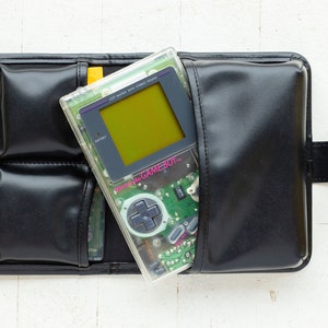 May include: A clear Game Boy Color handheld video game console with a green circuit board and a yellow screen. The console is in a black leather carrying case with multiple pockets.